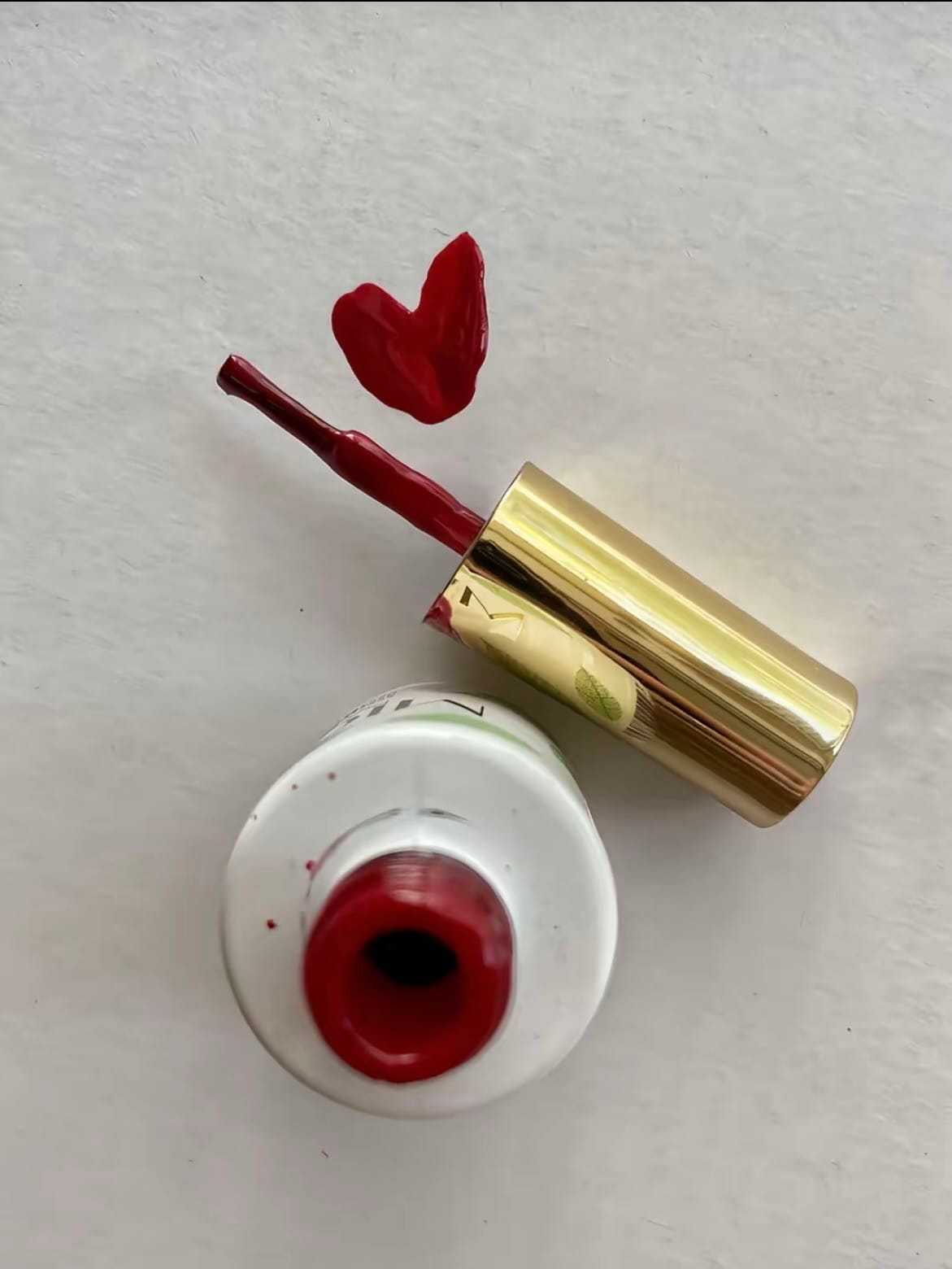 Open red nail polish bottle with a gold cap and heart-shaped brush strokes.